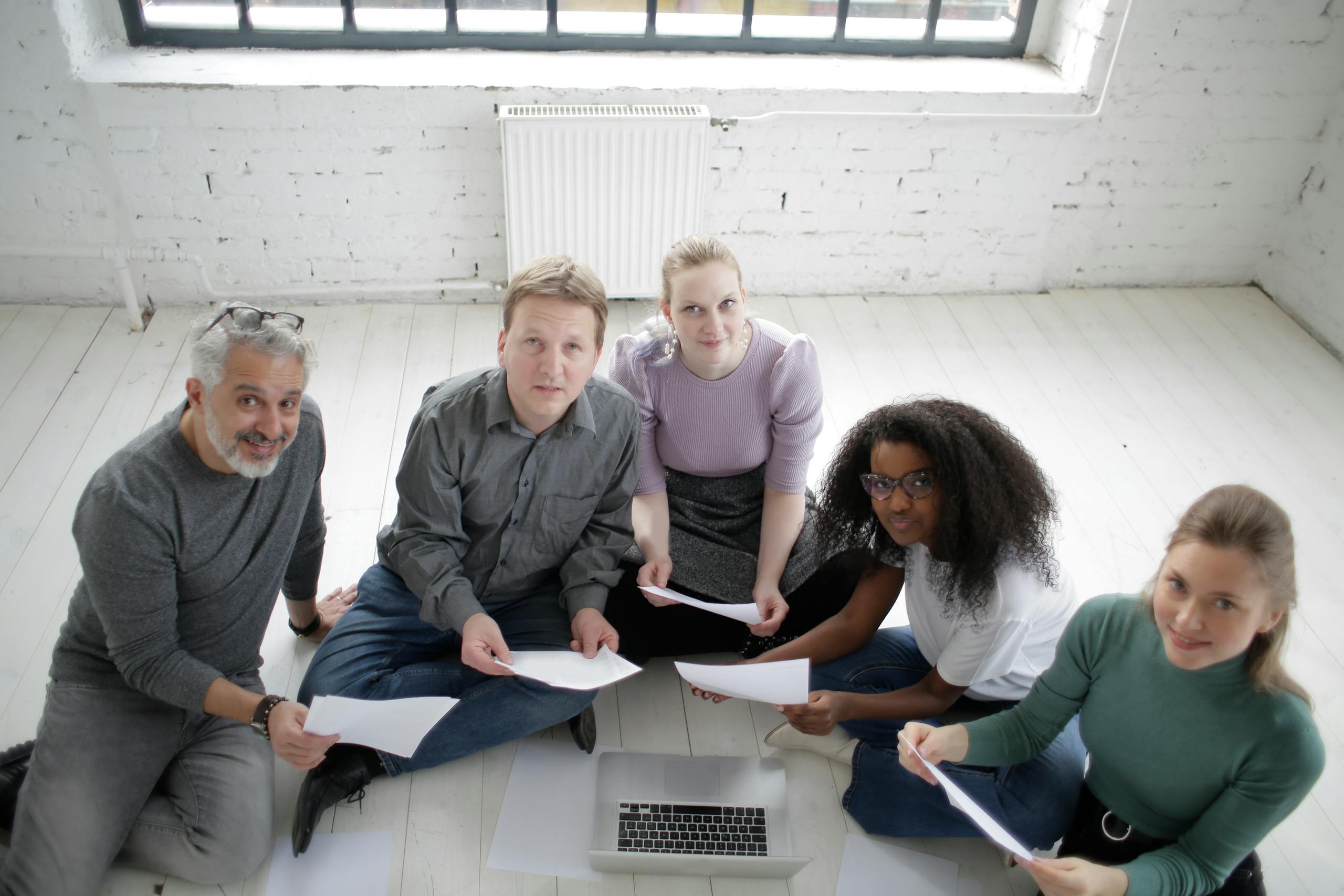 A diverse team of adults collaborating in a modern office, working on documents around a laptop.